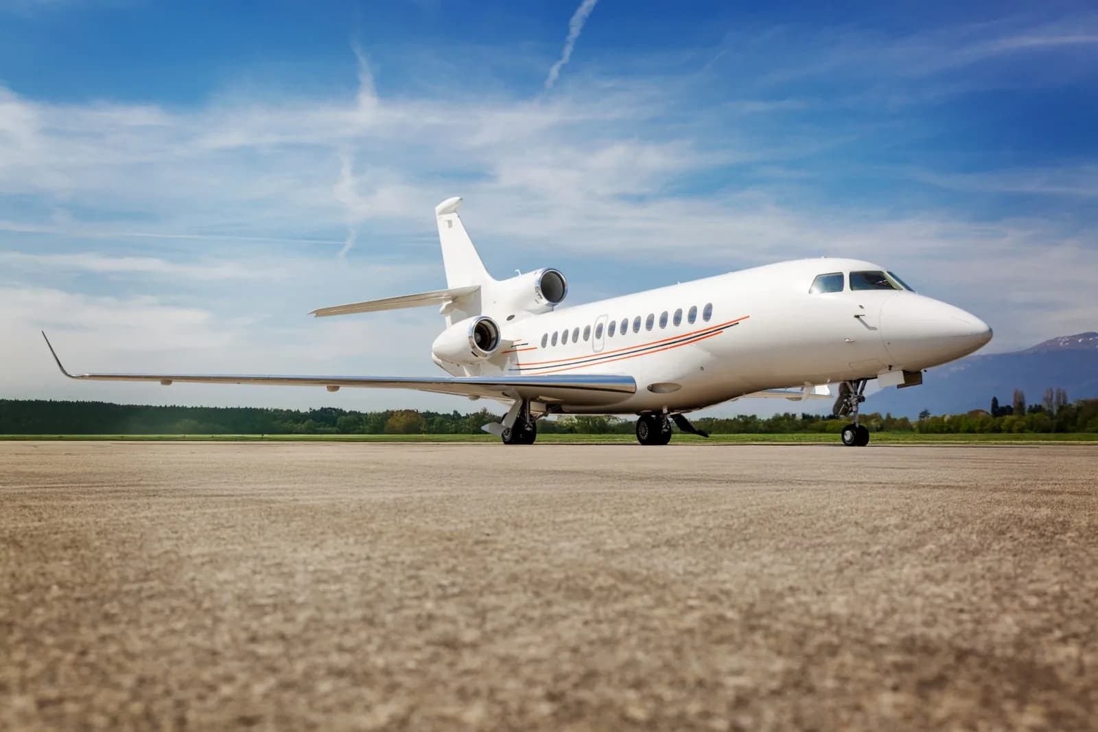 A white Dassault Falcon business jet with orange and red stripes on the fuselage, parked on an airfield under a blue sky.
