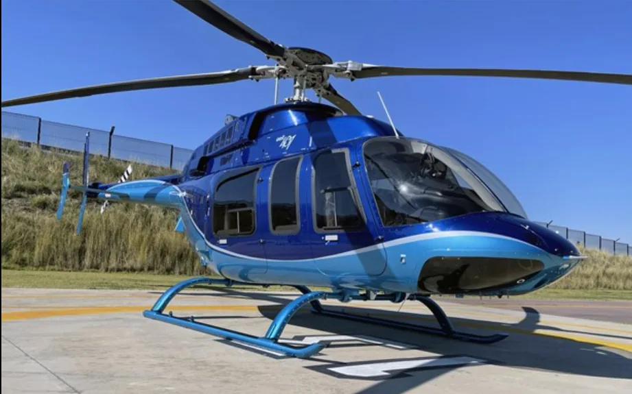 A shiny blue and light blue Bell 505 Jet Ranger X helicopter is parked on an airfield under a clear sky.