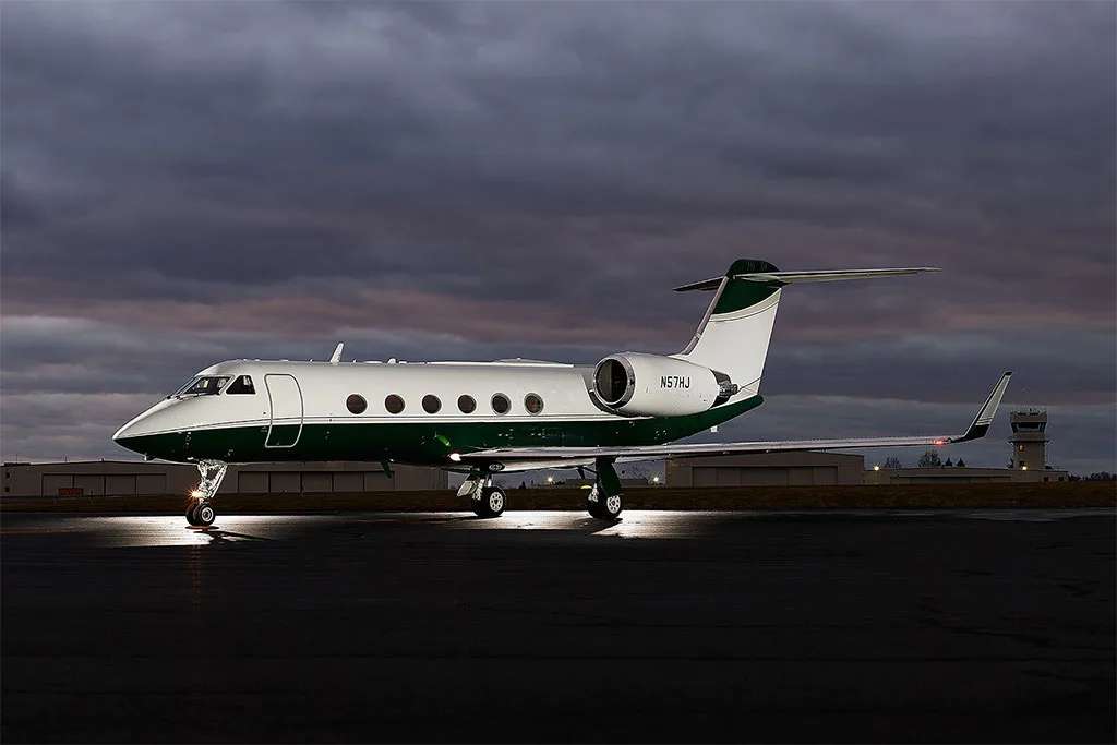 A white and dark green Gulfstream G550 aircraft parked on the tarmac at dusk with its landing lights on.