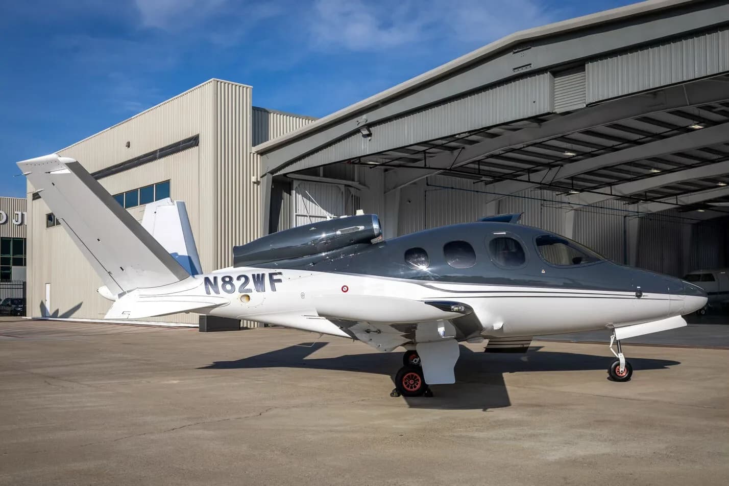 A three-quarter rear view of a white and dark grey Cirrus Vision Jet (N82WF) parked outside a hangar.