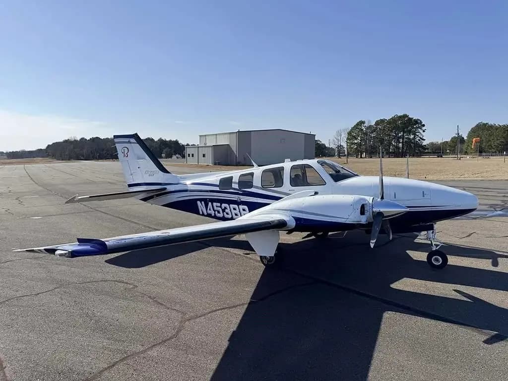 A white and blue Beechcraft Baron N453BB parked on an airfield with hangars in the background, viewed from the front-left.