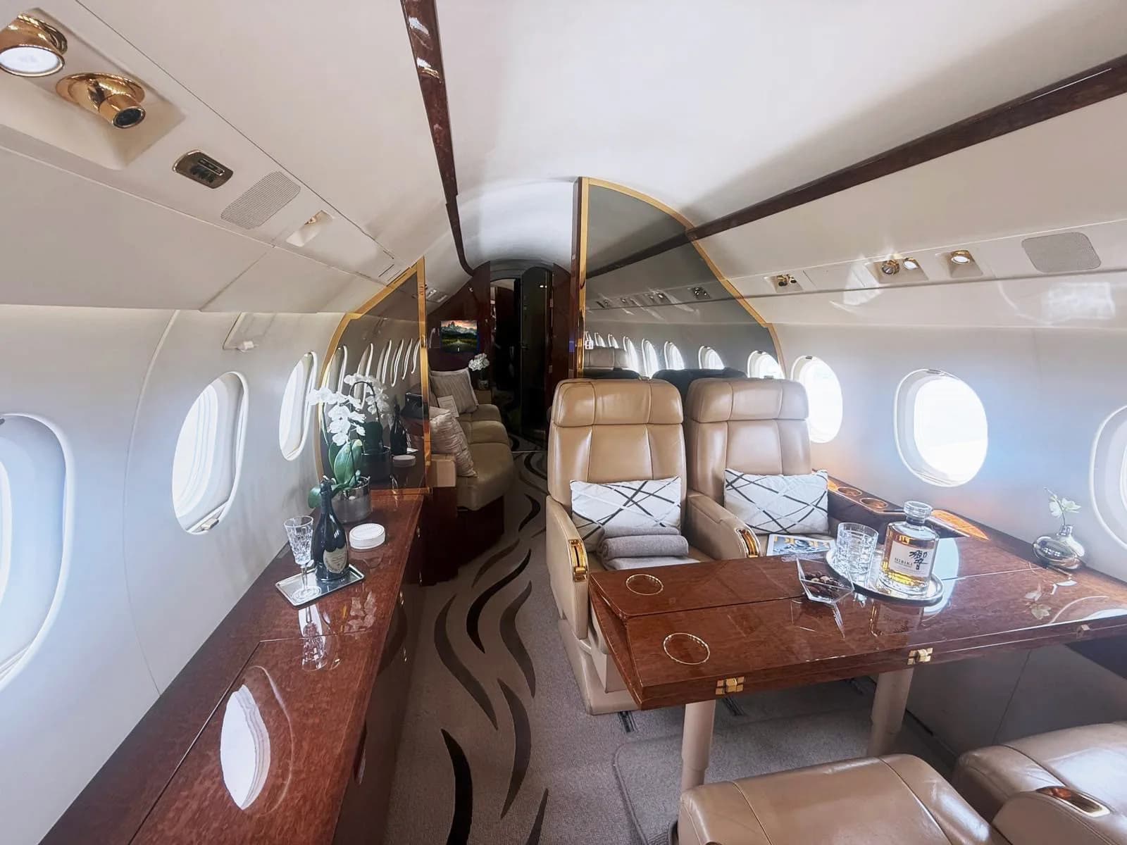 A luxurious aircraft cabin interior with tan leather seats, a polished wooden table set with drinks, and a patterned carpet.