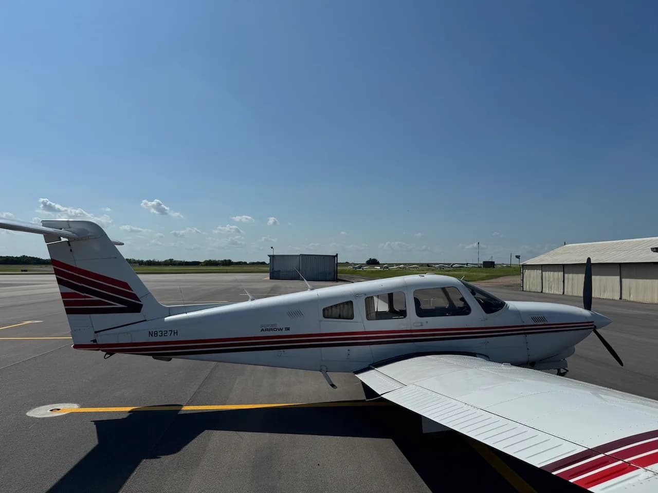 A white Piper Arrow II aircraft with red and brown stripes parked on an asphalt tarmac, viewed from the right side.
