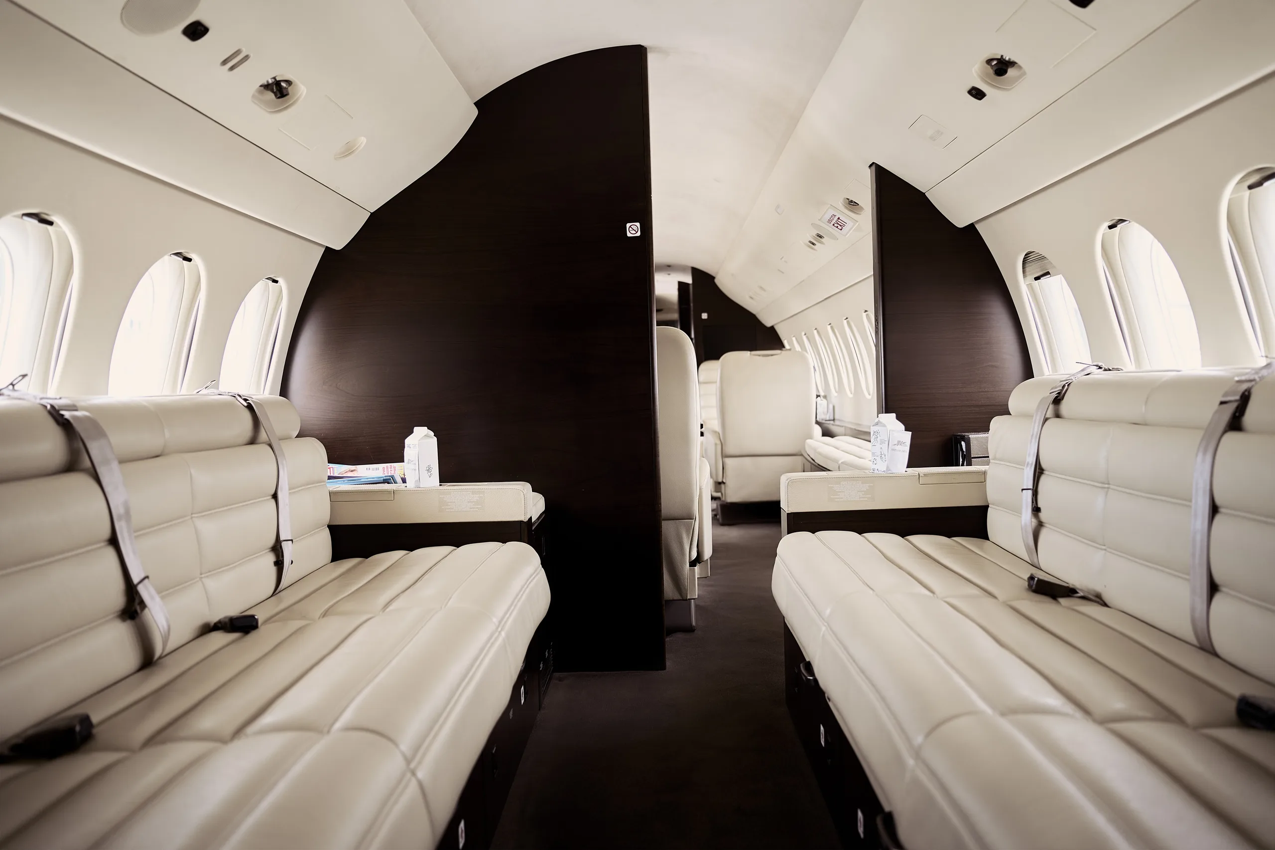 A view down the aisle of a private jet cabin, showing cream leather bench seating and a dark partition.
