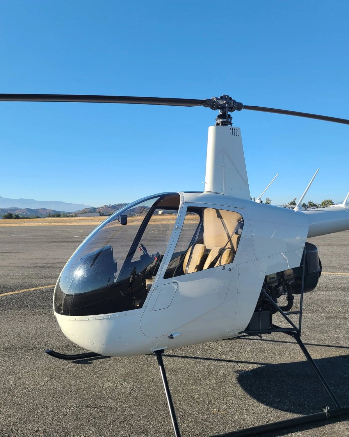 A white Robinson R22 helicopter is parked on an asphalt runway with mountains in the background.