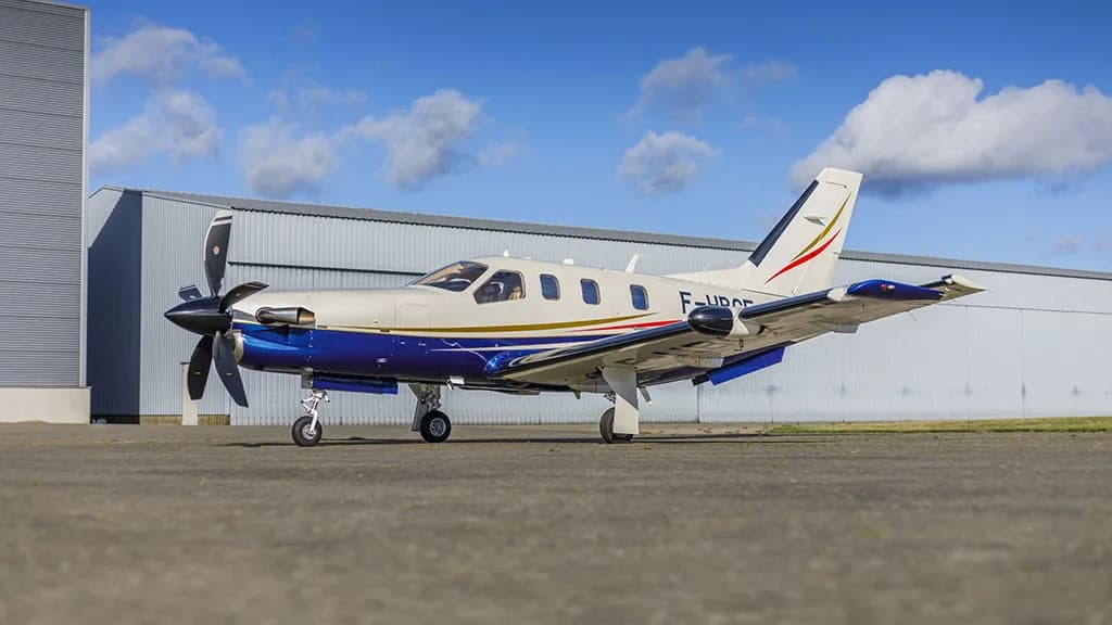 A white and blue TBM aircraft with registration F-HBGE parked on an airfield with a large hangar in the background.