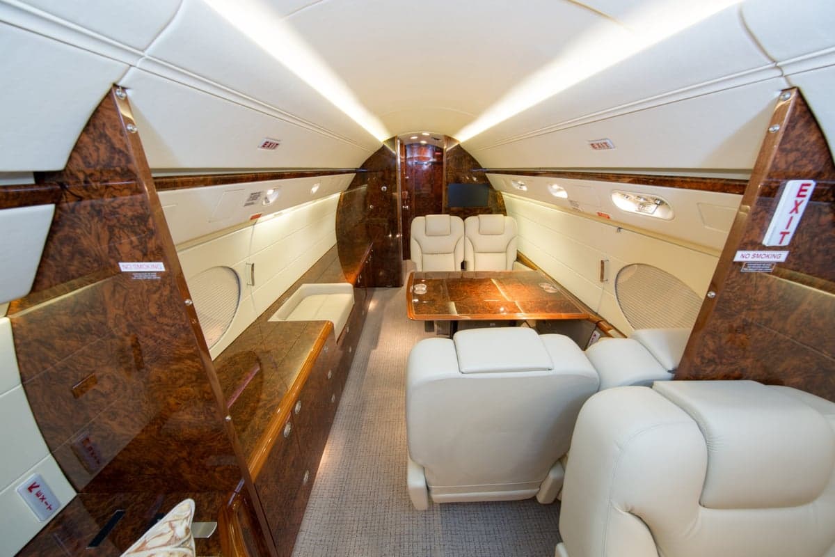 The interior of a private jet cabin featuring a dining table, cream leather seats, and rich burl wood cabinetry.