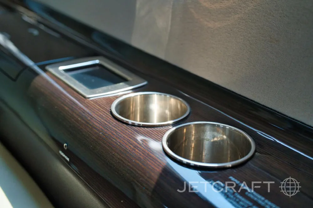 A close-up of two silver cup holders and a control panel set into a dark wood-grain side panel in an aircraft cabin.