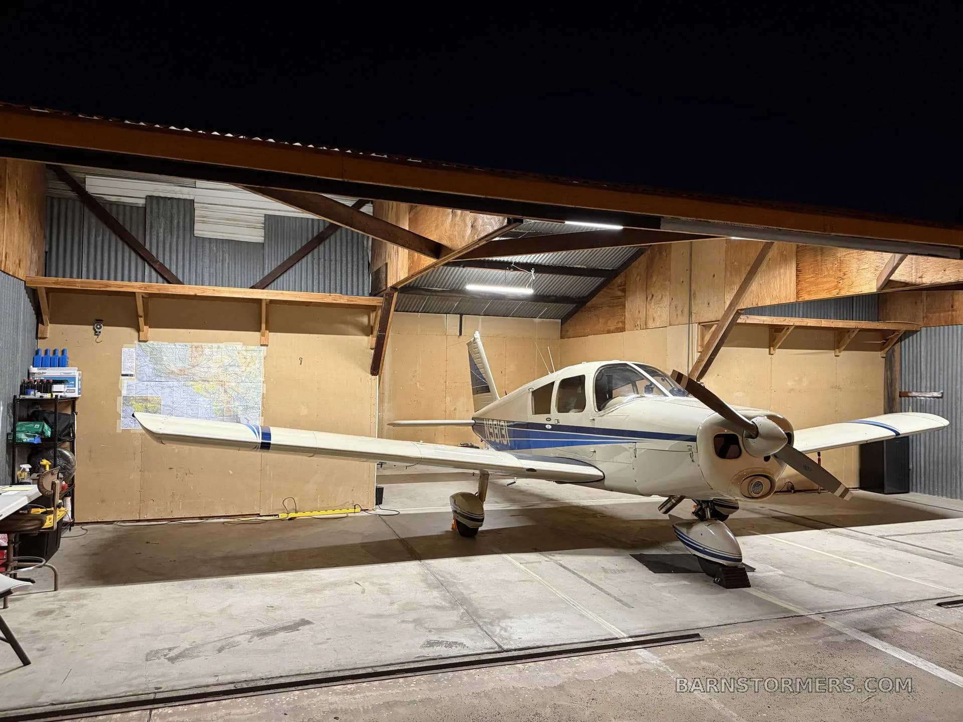 A white and blue single-engine propeller aircraft (N9813I) is parked in a hangar at night, illuminated by overhead lights.