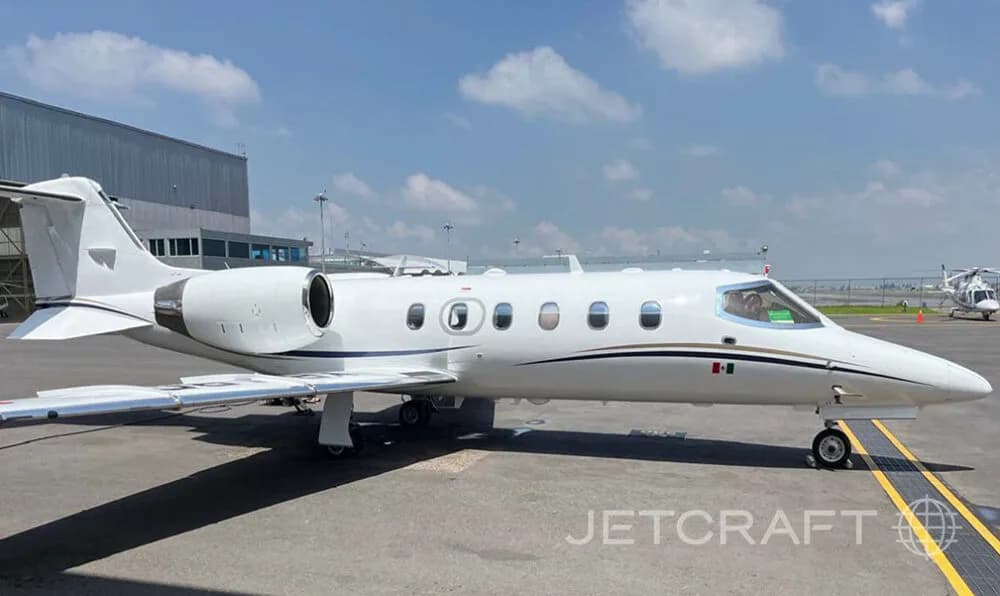 A white business jet with a black and gold stripe and a Mexican flag on the nose, parked on an airfield next to a hangar.