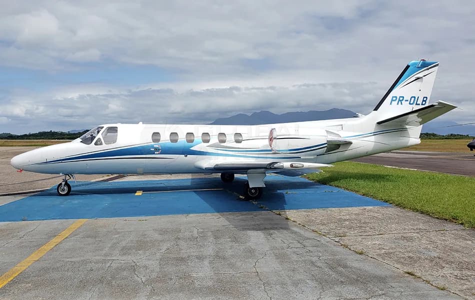 A white business jet with blue and black stripes, registration PR-OLB, parked on an airfield with mountains in the background.