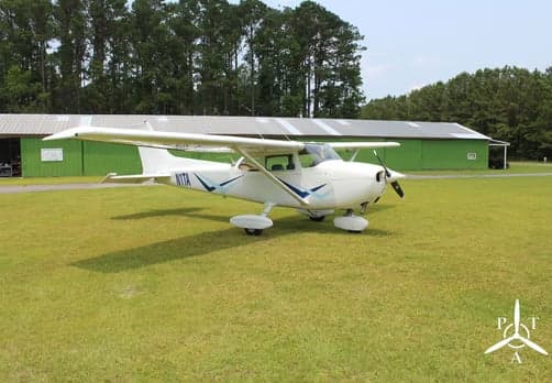 A white Cessna 172 aircraft with blue stripes and tail number N1TA is parked on a grassy airfield in front of green hangars.