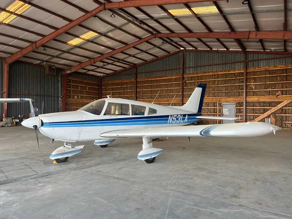 A white Piper Cherokee aircraft with blue stripes, registration N53LA, parked inside a hangar.