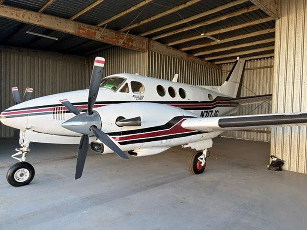 A white and red Beechcraft King Air C90 aircraft with registration N717JG parked in a hangar, viewed from the front-left.