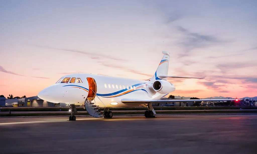 A white private jet with blue and orange stripes on the fuselage and tail, parked on an airfield at sunset with its main door open.