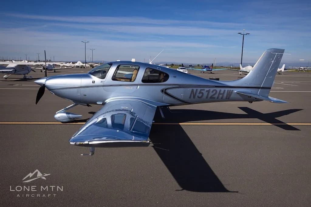 Side view of a silver Cirrus SR22 aircraft, N512HW, parked on an airfield with other planes in the background.