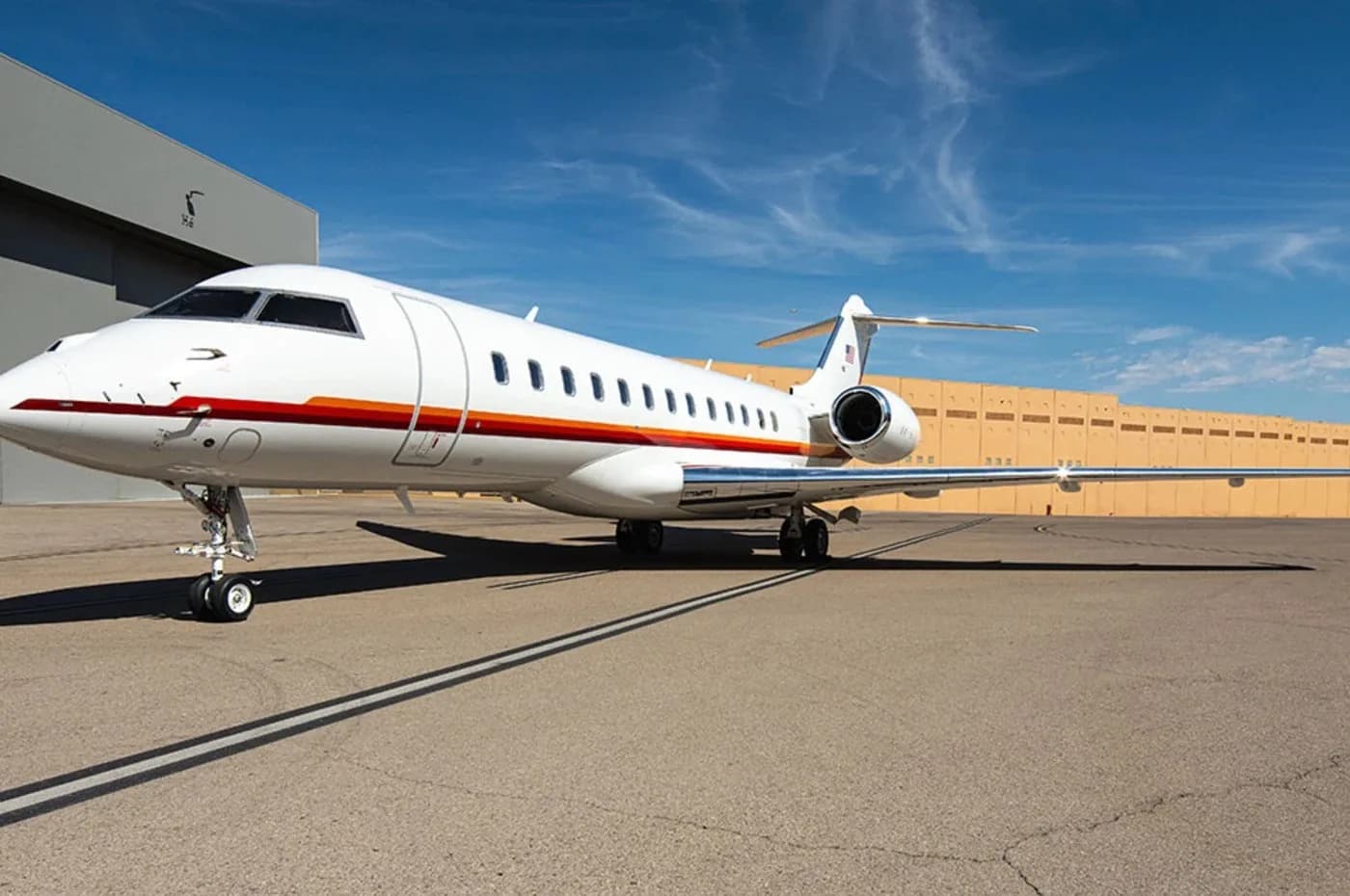 A white Bombardier Global business jet with red and orange stripes on the fuselage is parked on an airfield.