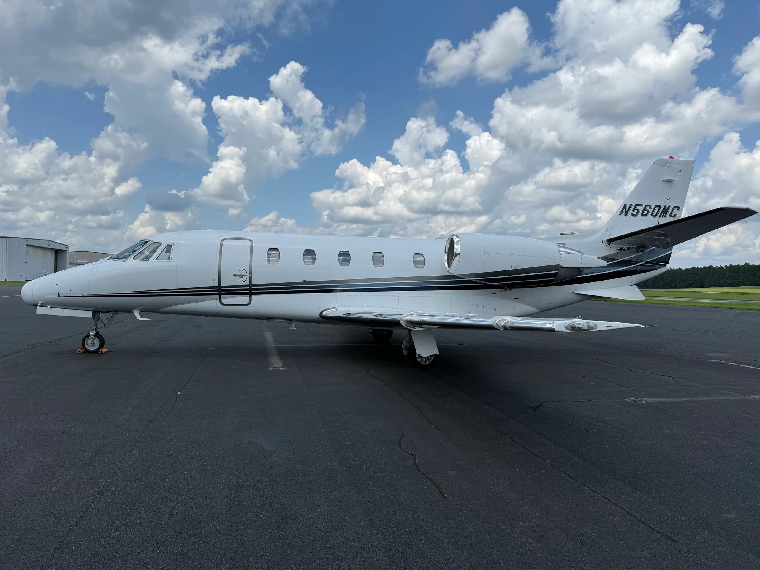 A white Cessna 560XL Citation XLS business jet with black and grey stripes is parked on an asphalt apron under a cloudy blue sky.