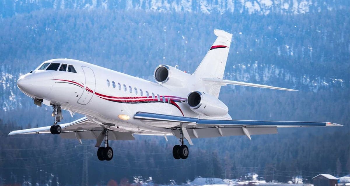 A white business jet with red and black stripes on its fuselage, landing or taking off against a snowy, forested mountain background.