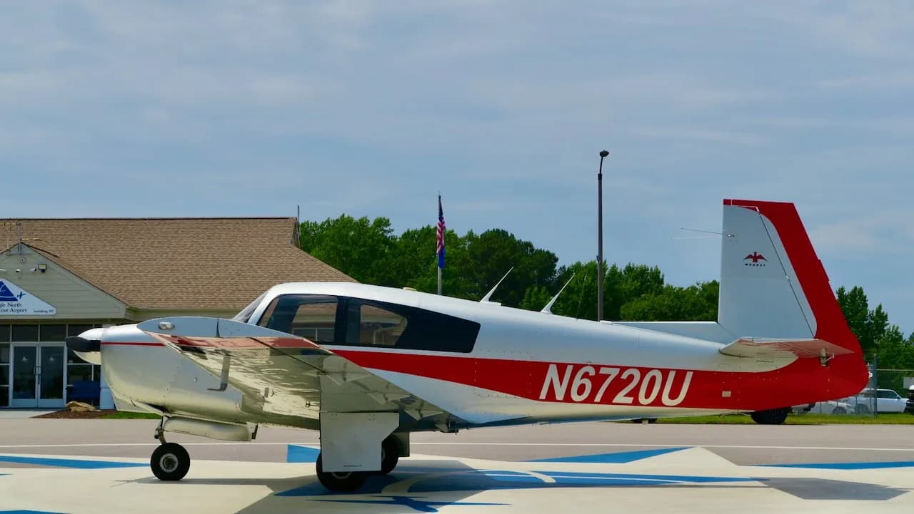 Left side view of a silver and red Mooney aircraft with registration N6720U parked in front of an airport building.