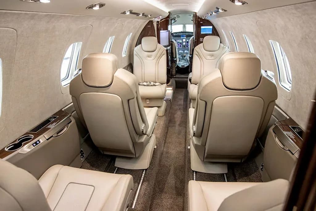 The luxurious interior cabin of a private jet features light tan leather seats arranged in a club configuration, looking towards the cockpit.