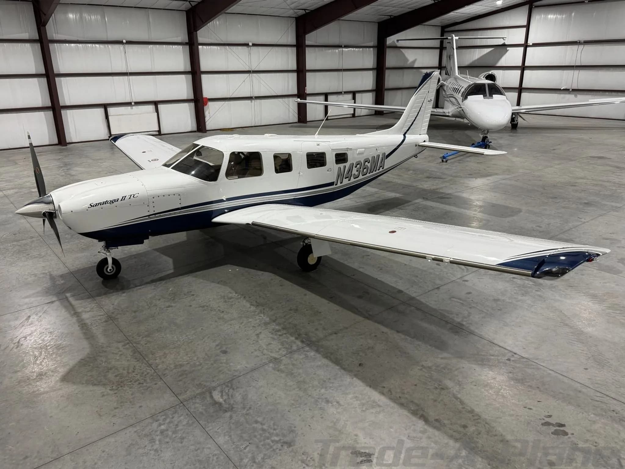 A white Piper Saratoga II TC N436MA with blue stripes is parked in a hangar with other aircraft.