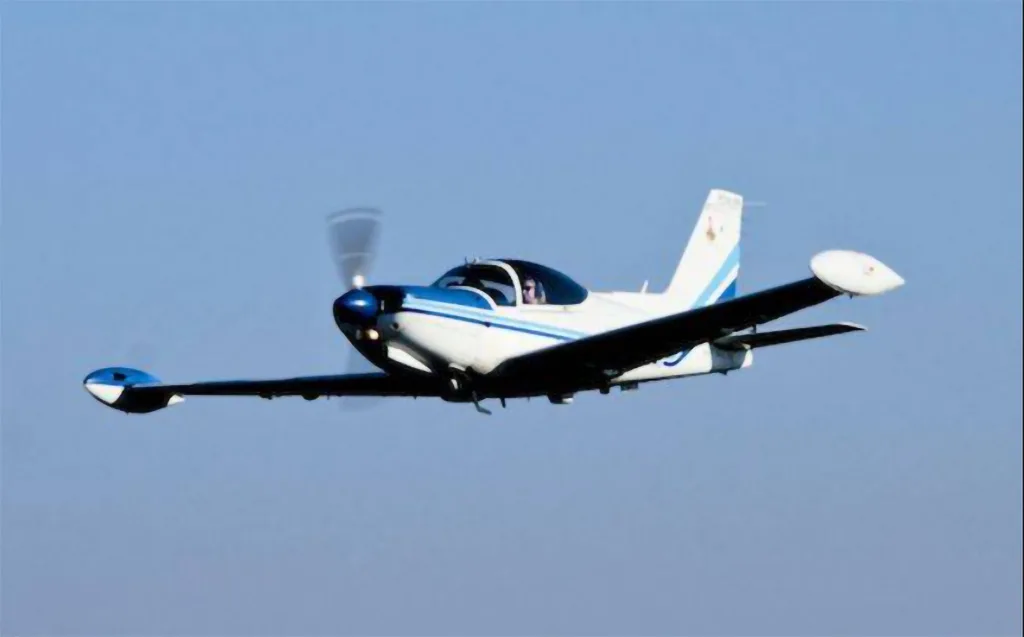 A white and blue single-engine aircraft with wingtip tanks is flying against a clear blue sky.