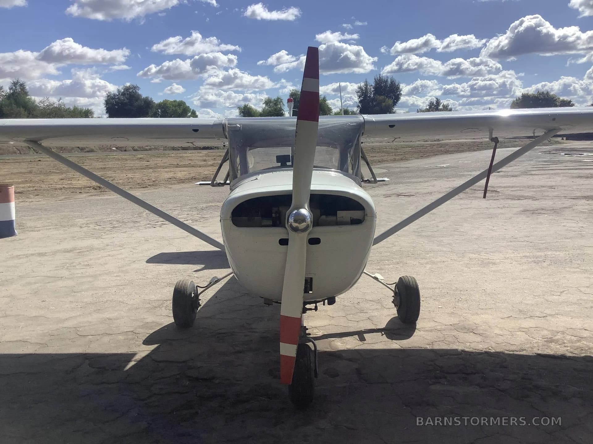 A front view of a white Cessna 150 with a red-tipped propeller, parked on a cracked asphalt surface under a blue sky with white clouds.
