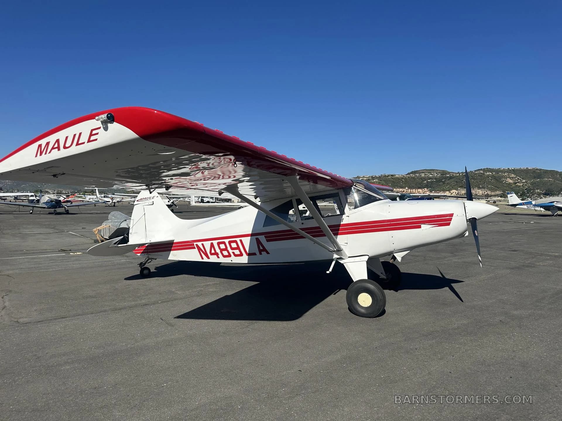 A white Maule M-7-235 aircraft with red stripes and registration N489LA is parked on an asphalt tarmac with hills in the background.