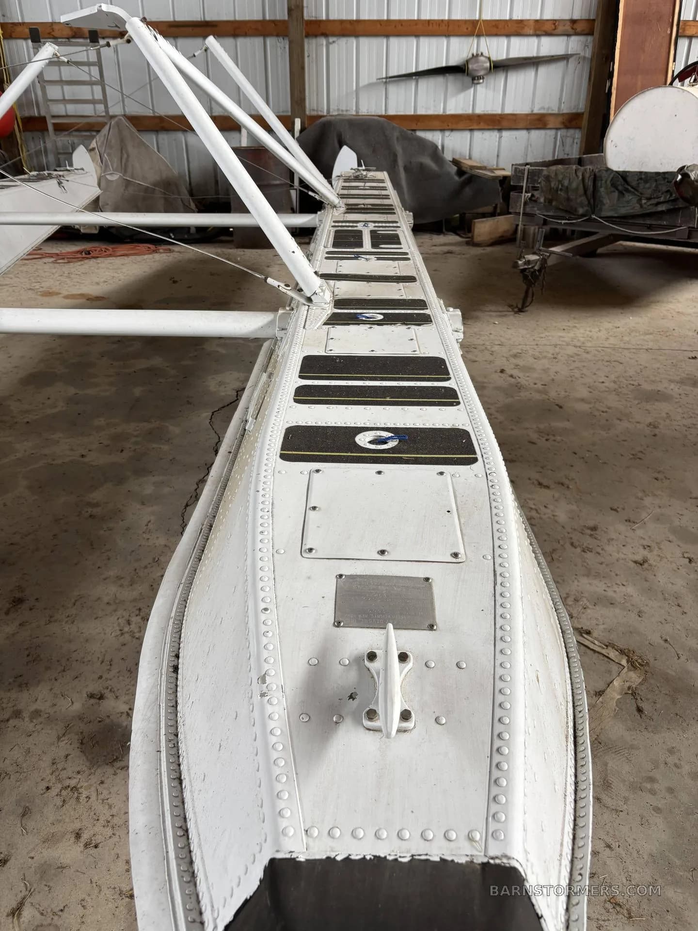 An overhead view of a white aircraft float with black anti-slip pads and a mooring cleat, showing its long, riveted structure in a hangar.