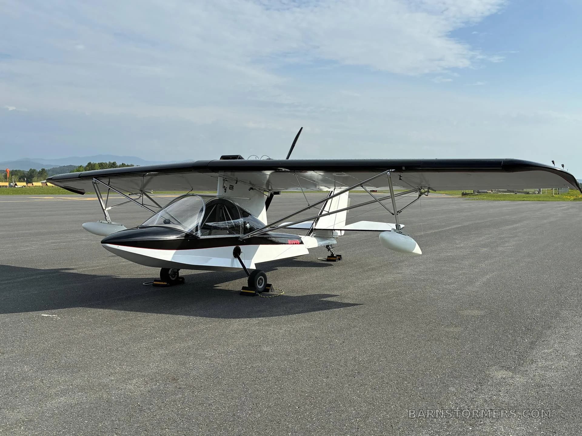 A white and black ultralight aircraft with registration N7749D parked on an airfield with mountains in the background under a partly cloudy sky.