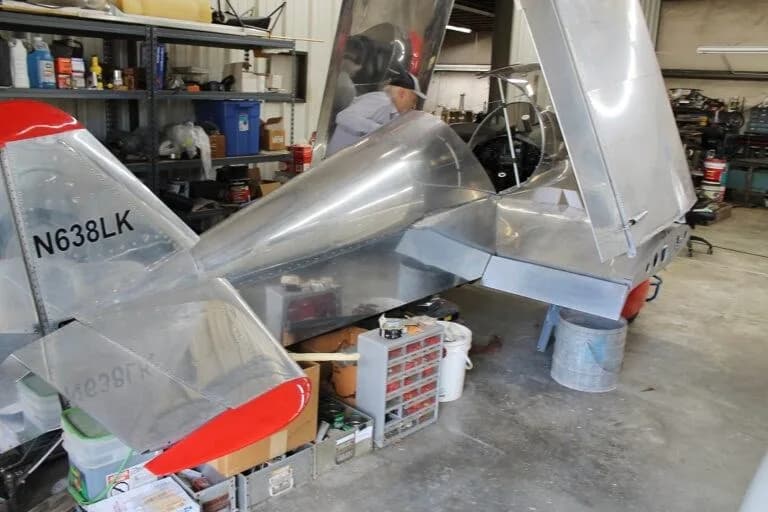 The tail of a silver experimental aircraft with red accents and registration N638LK is visible in a workshop, with a person working inside the cockpit.