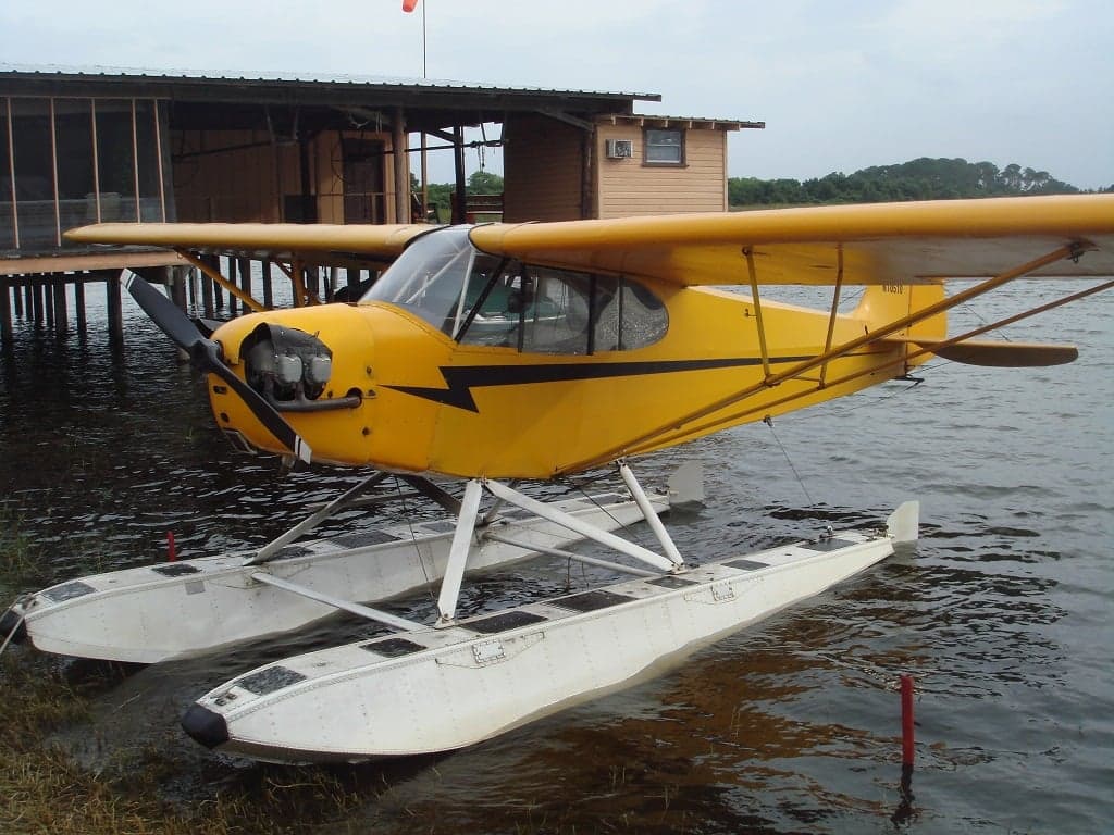 A yellow Piper Cub on floats is docked near a wooden building.
