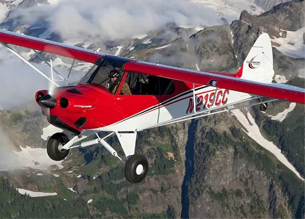 A red and white Carbon Cub aircraft flying near mountains.
