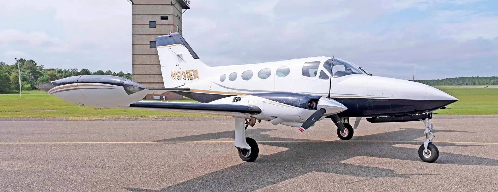 A white and dark blue Cessna 414 Chancellor with gold stripes, registration N991EM, parked on an airfield with a control tower in the background.