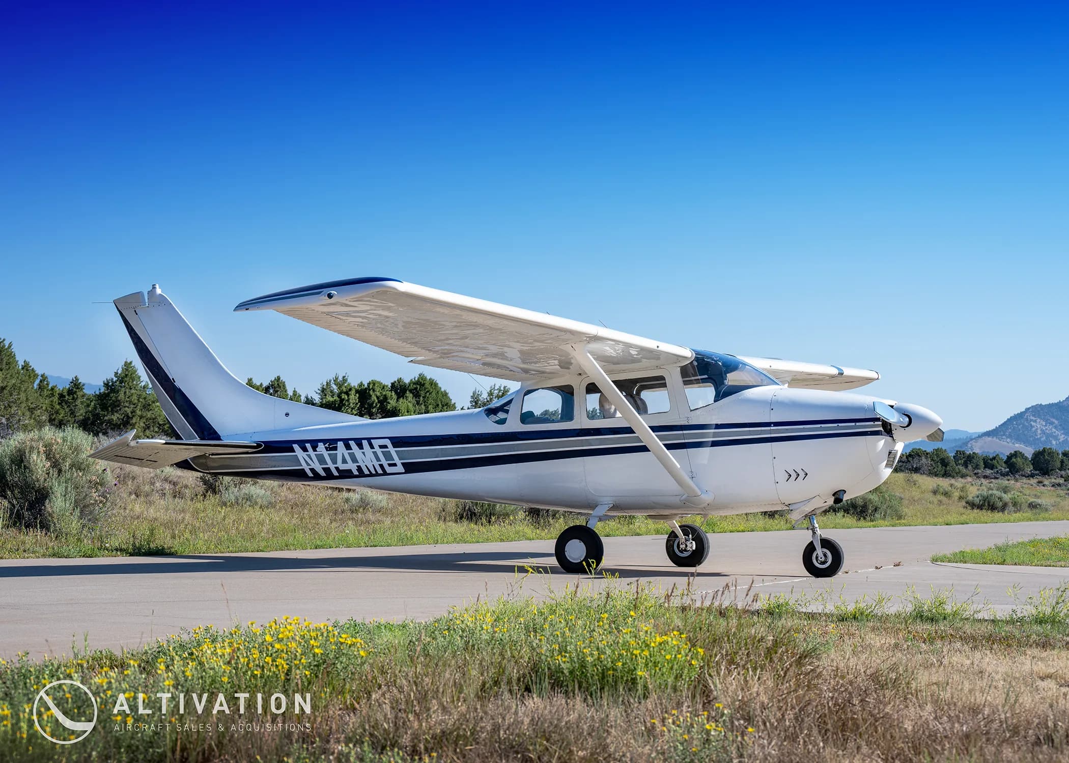 A white Cessna 182 with blue and grey stripes, registration N74MD, parked on a paved surface with a mountain in the background.