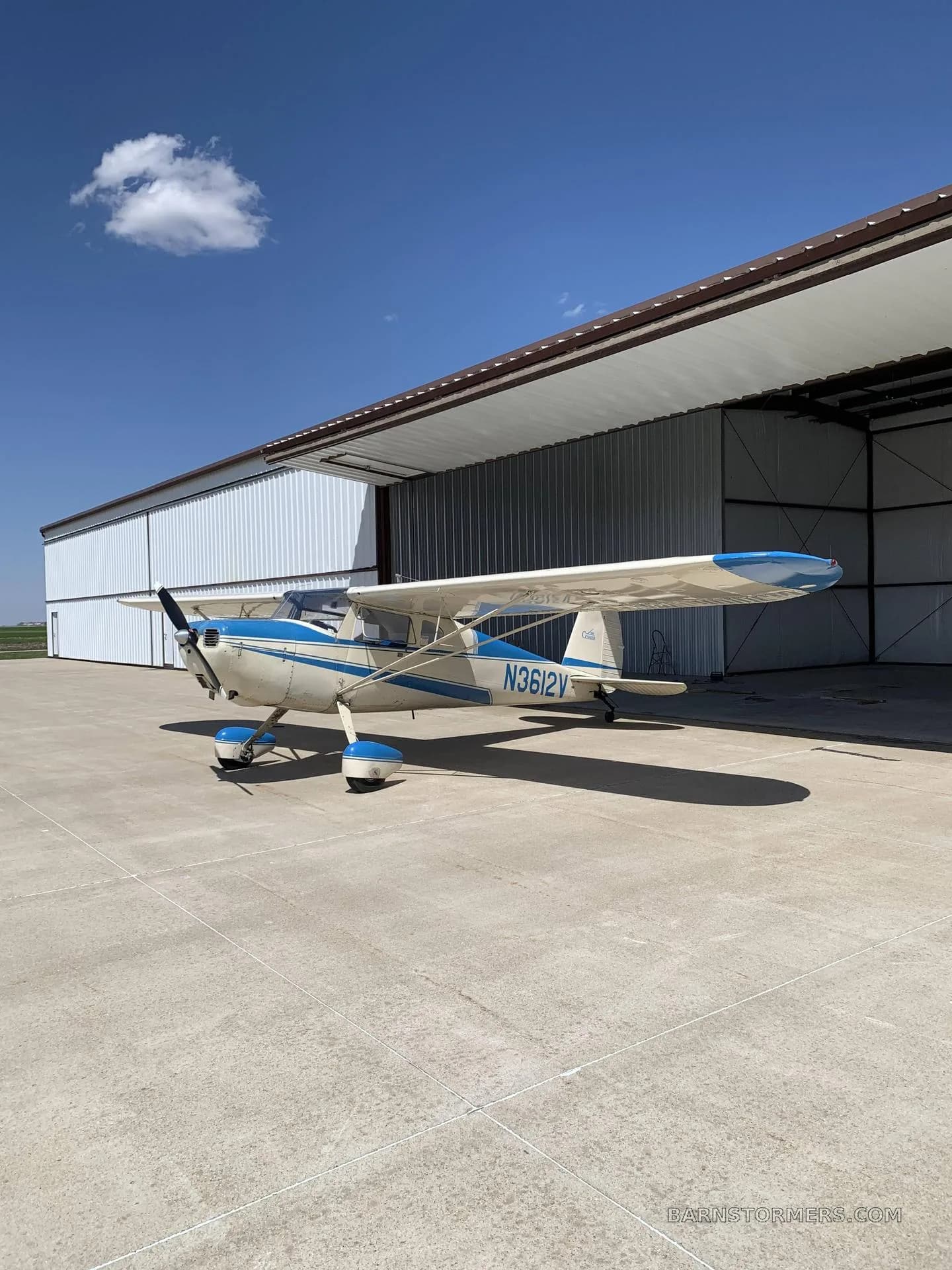 A white and blue Cessna 170B aircraft with tail number N3612V parked on a concrete pad outside a hangar under a clear blue sky.