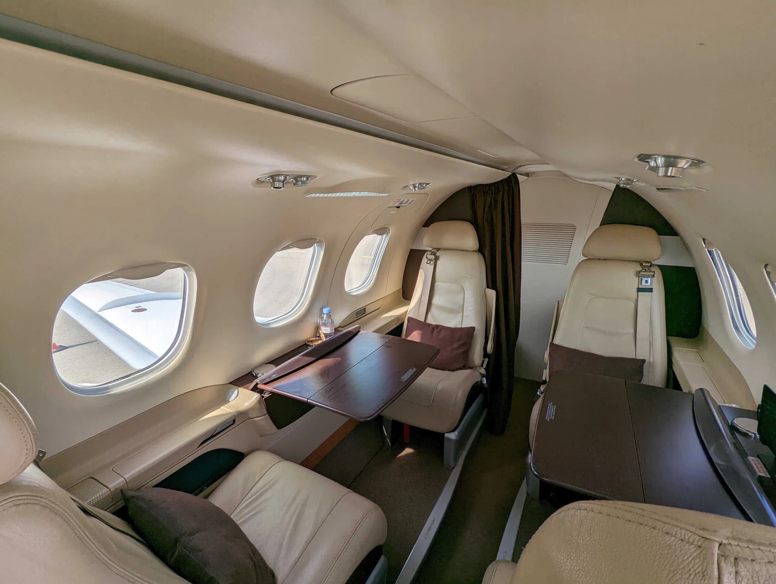 A view inside a private jet cabin shows beige leather seats, fold-out tables, and windows looking out onto an aircraft wing.