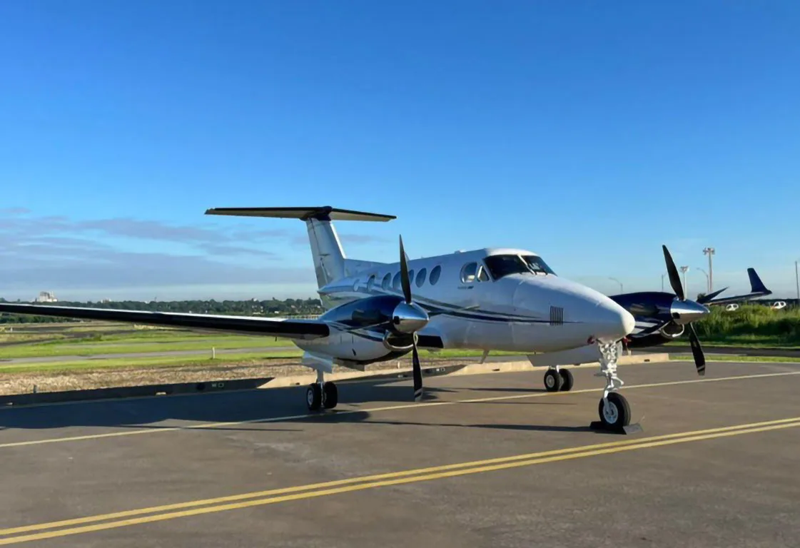 A white and dark blue twin-turboprop aircraft parked on an airport tarmac with another aircraft in the background.