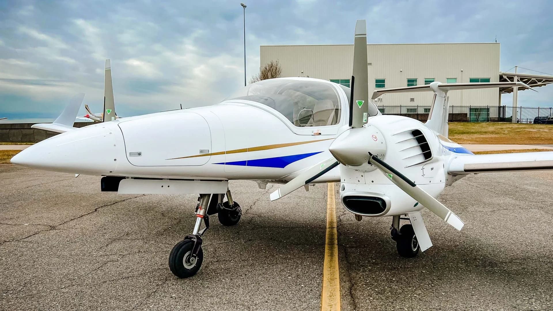 A white Diamond DA42 twin-engine aircraft with blue and gold stripes is parked on an asphalt tarmac with a building in the background.