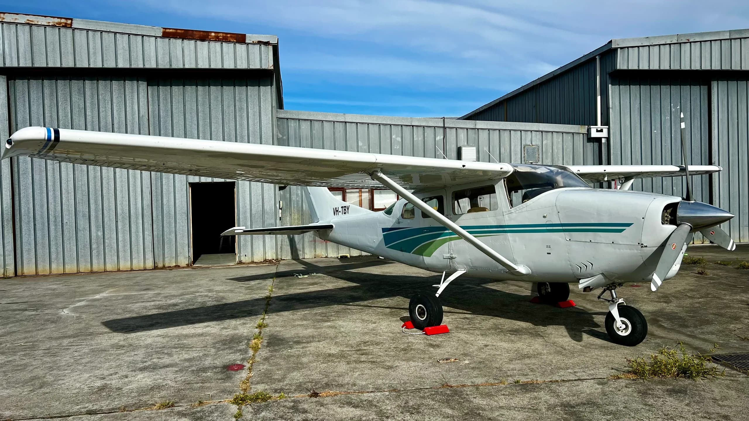 A white single-engine propeller aircraft with blue and green stripes parked on concrete next to metal hangars.