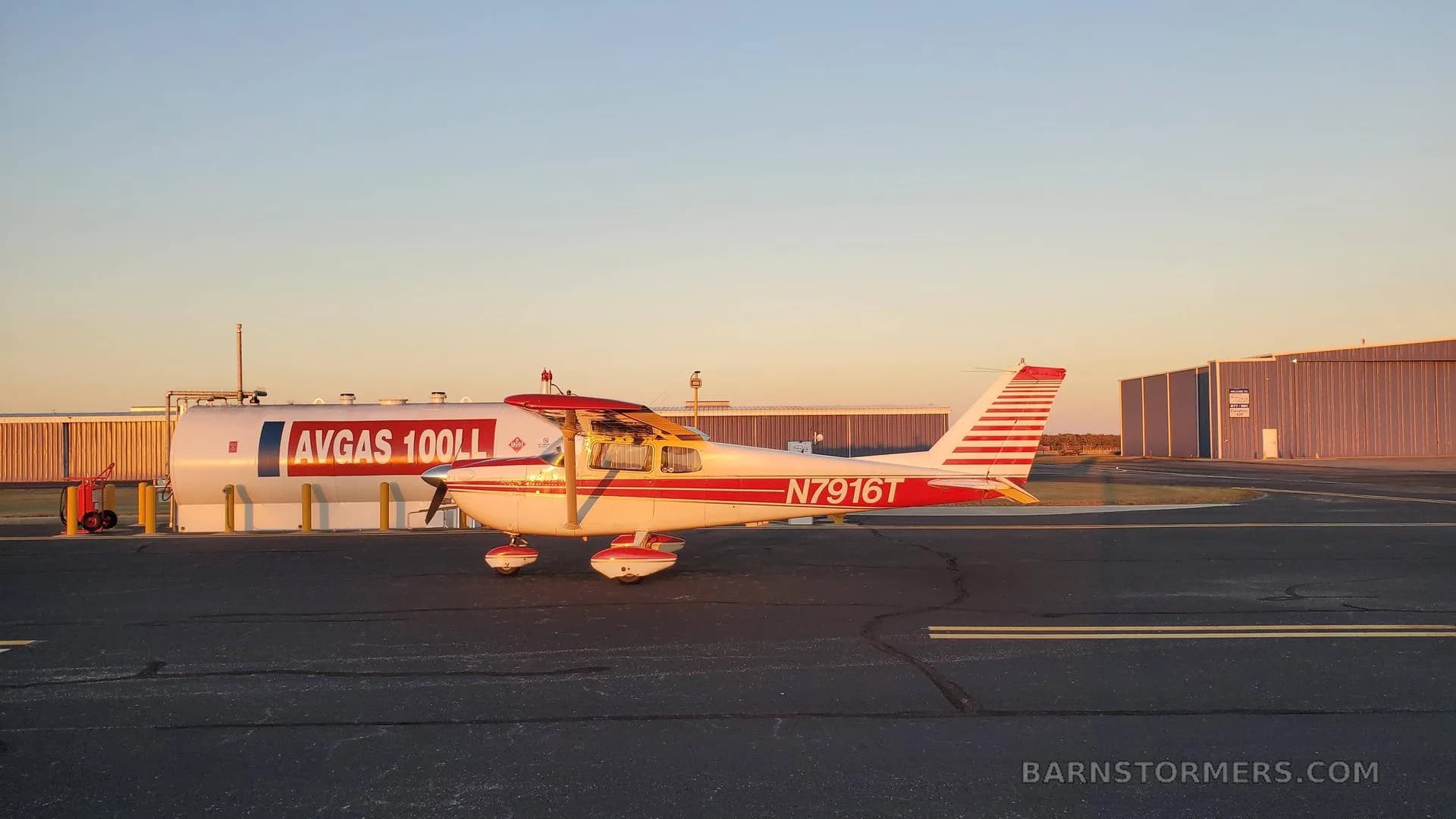 A white and red Cessna 172 with registration N7916T parked next to an AVGAS fuel tank at sunset.
