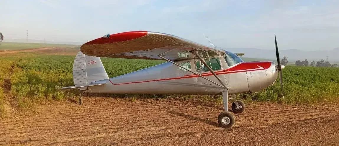 A silver and red Cessna 170 aircraft is parked on a dirt field.