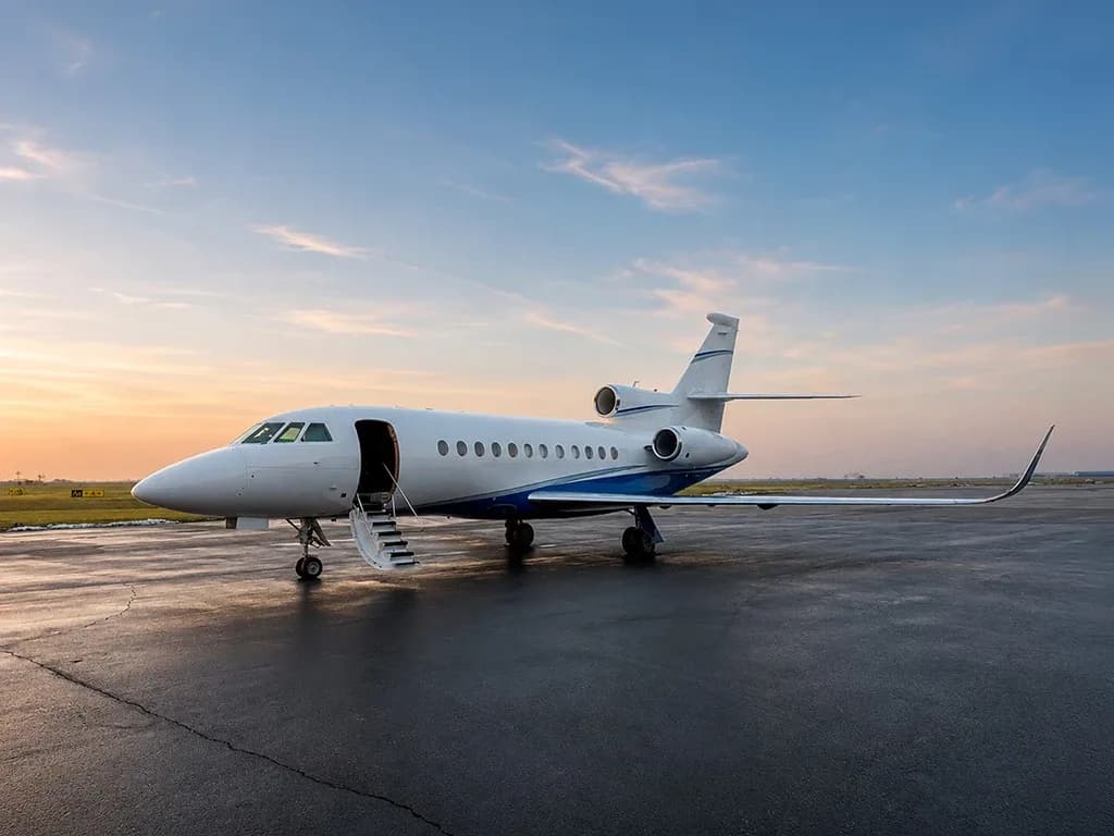 A white business jet with blue accents and an open entry door parked on a wet tarmac at sunset.