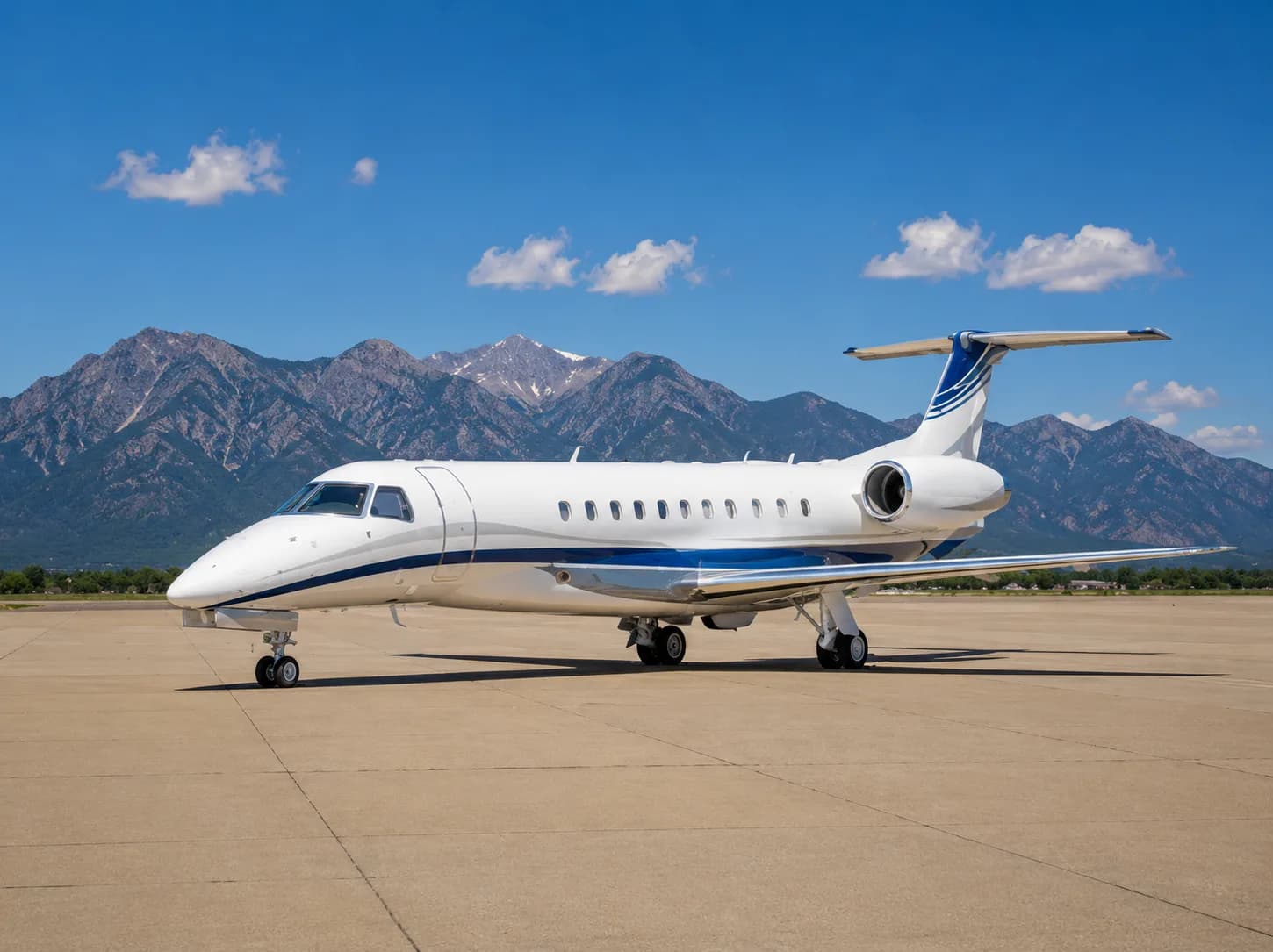 A white business jet with blue accent stripes is parked on an airfield with mountains in the background under a clear blue sky.