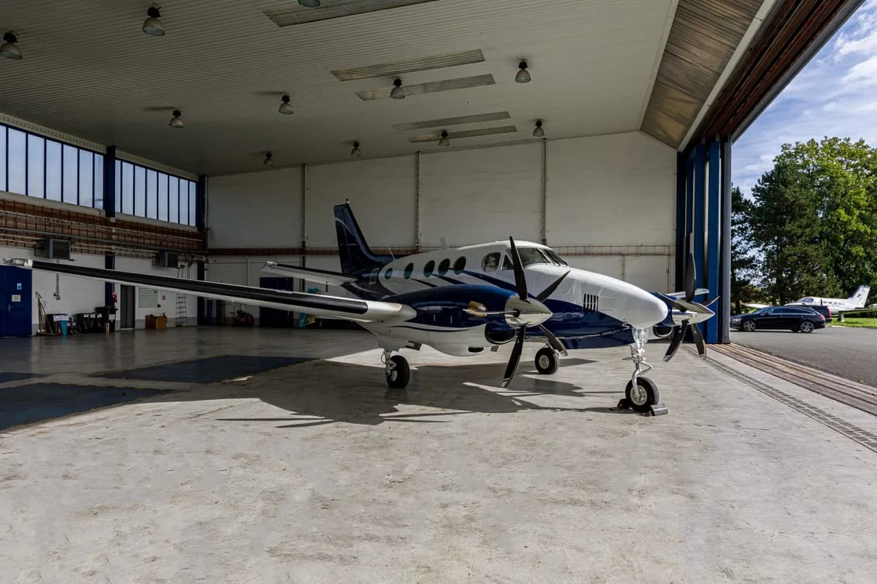 A white and blue Beechcraft King Air C90 twin-turboprop aircraft parked inside a hangar, with a car visible outside.