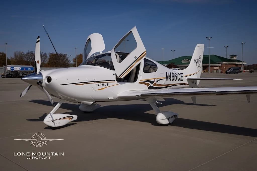 A white Cirrus SR22 aircraft with gold and black stripes, registration N466CE, parked on an airfield with its doors open.