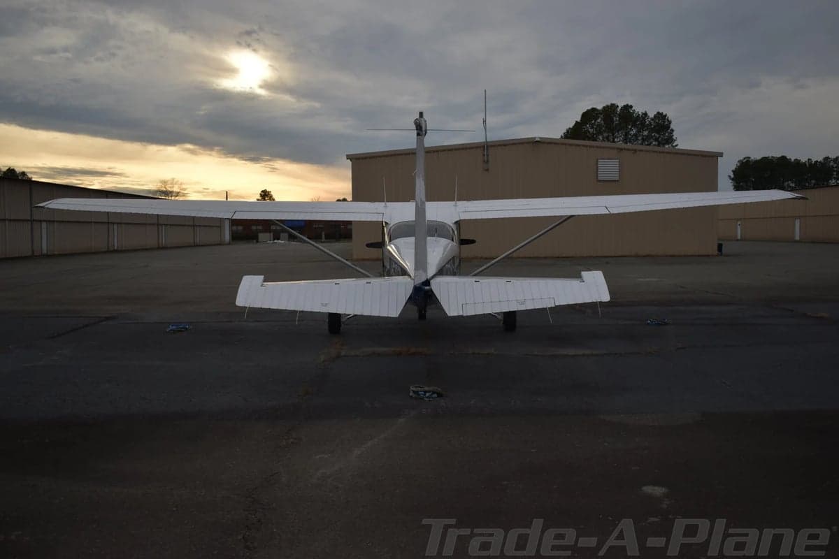 The rear view of a white Cessna 172 is shown on a tarmac at sunset, with a building in the background.