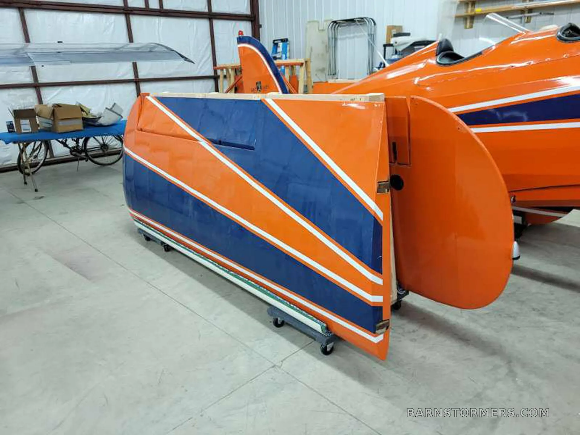 A close-up view of a disassembled orange and blue aircraft wing section on a dolly in a workshop.
