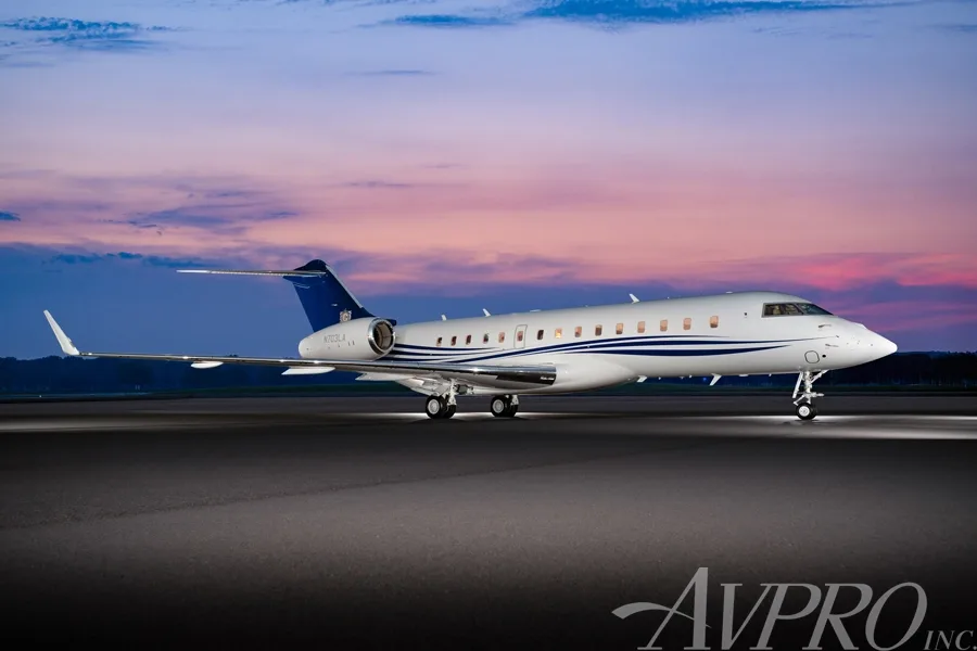 A Bombardier Global 6000 business jet with blue and white livery on a tarmac at sunset.